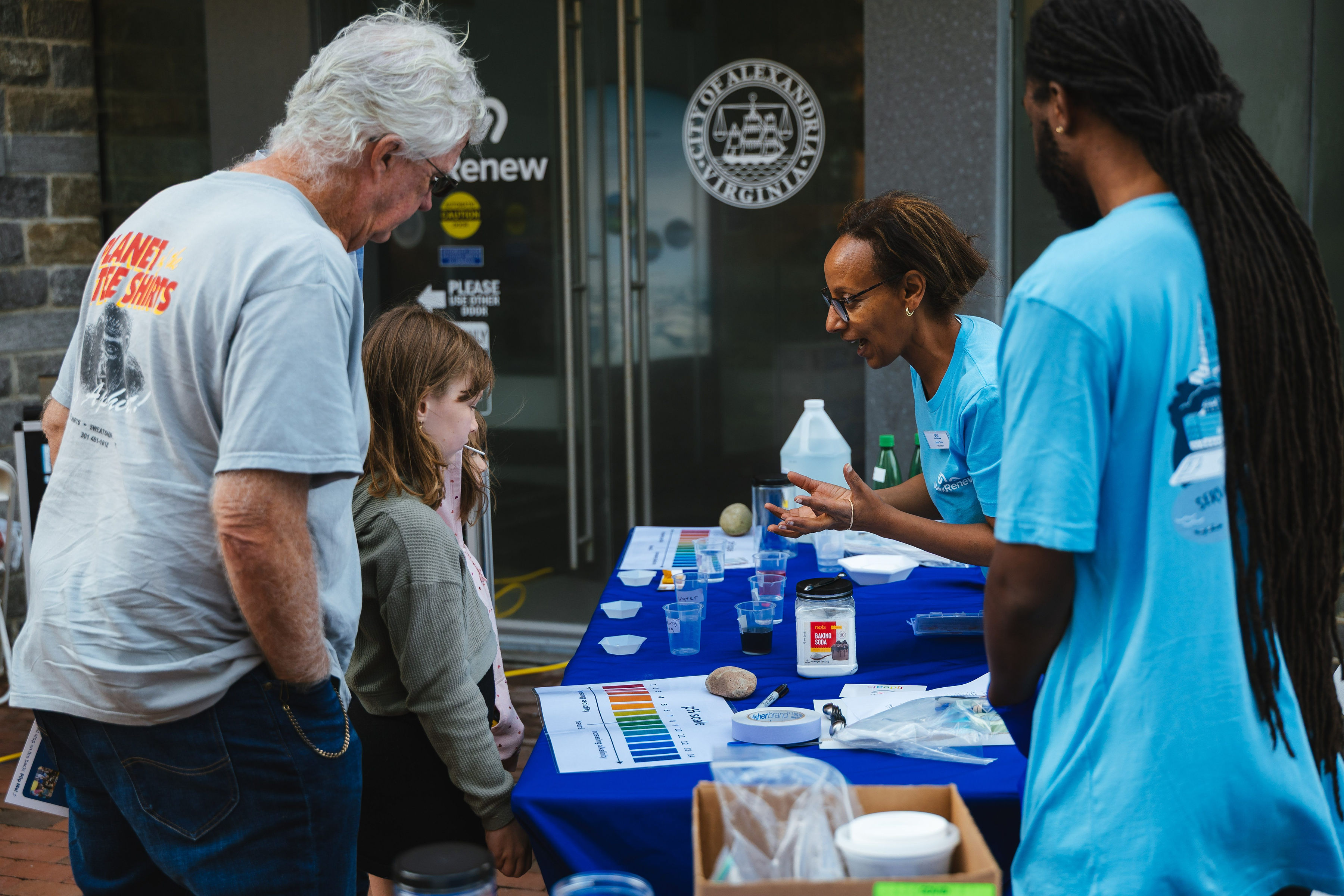 A woman in a blue AlexRenew t-shirt explains an activity involving pH and different solutions to a child and adult.