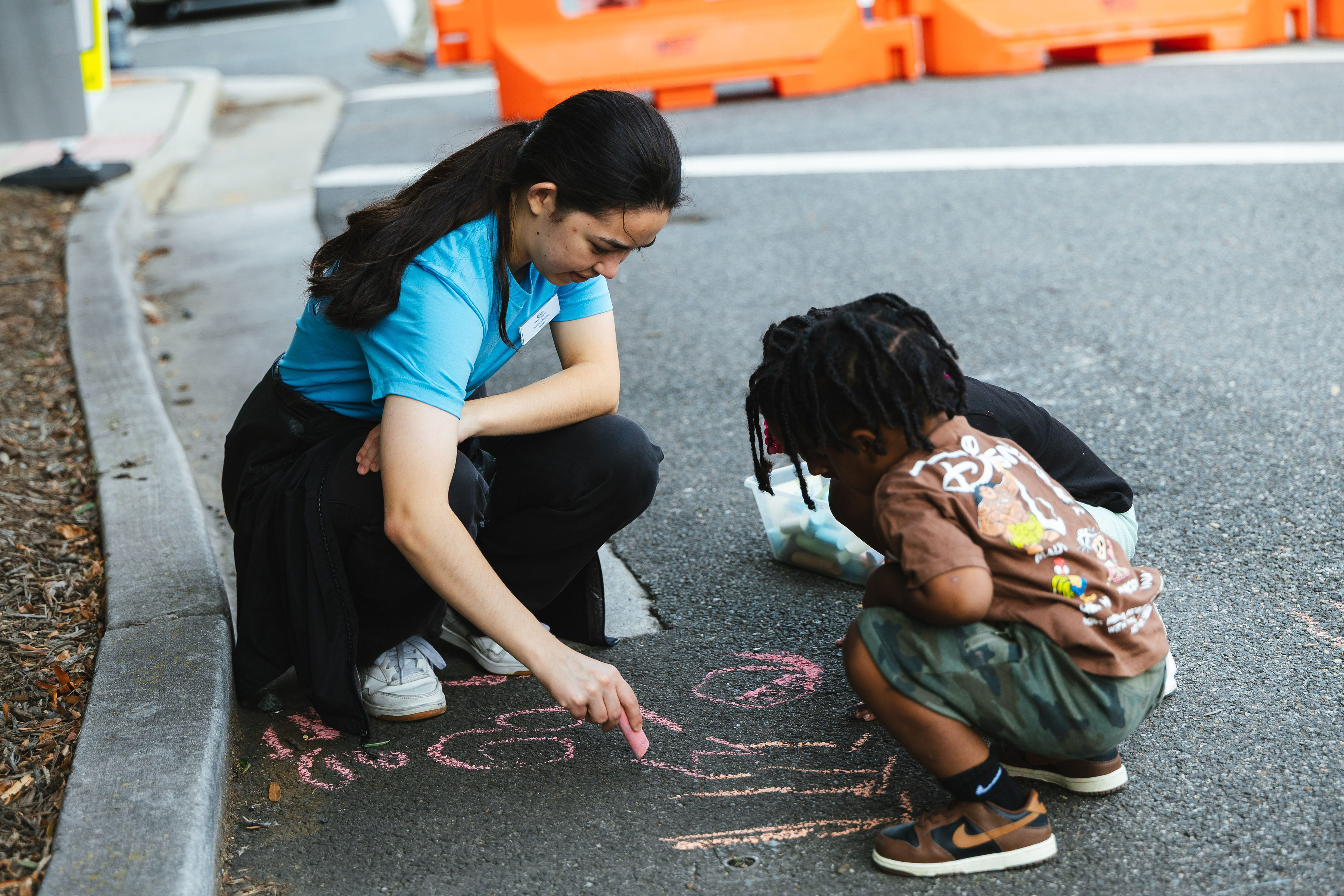 A woman with dark hair in a blue AlexRenew shirt crouches on the ground next to two children. All three people draw on the asphalt with sidewalk chalk.
