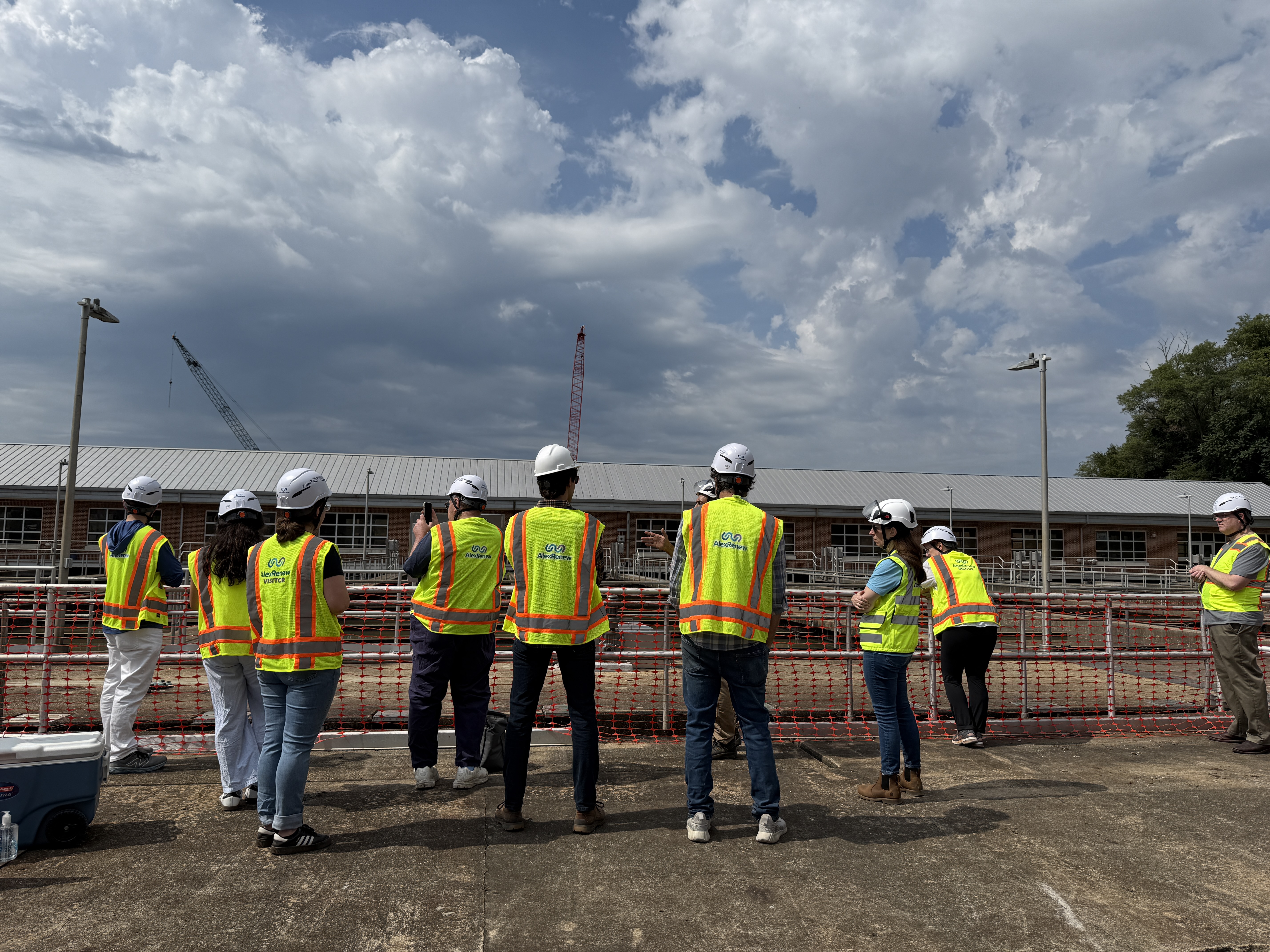 Photo of several people in hard hats and high viz vests from the back, as they listen to a tour leader talk about treatment tanks.