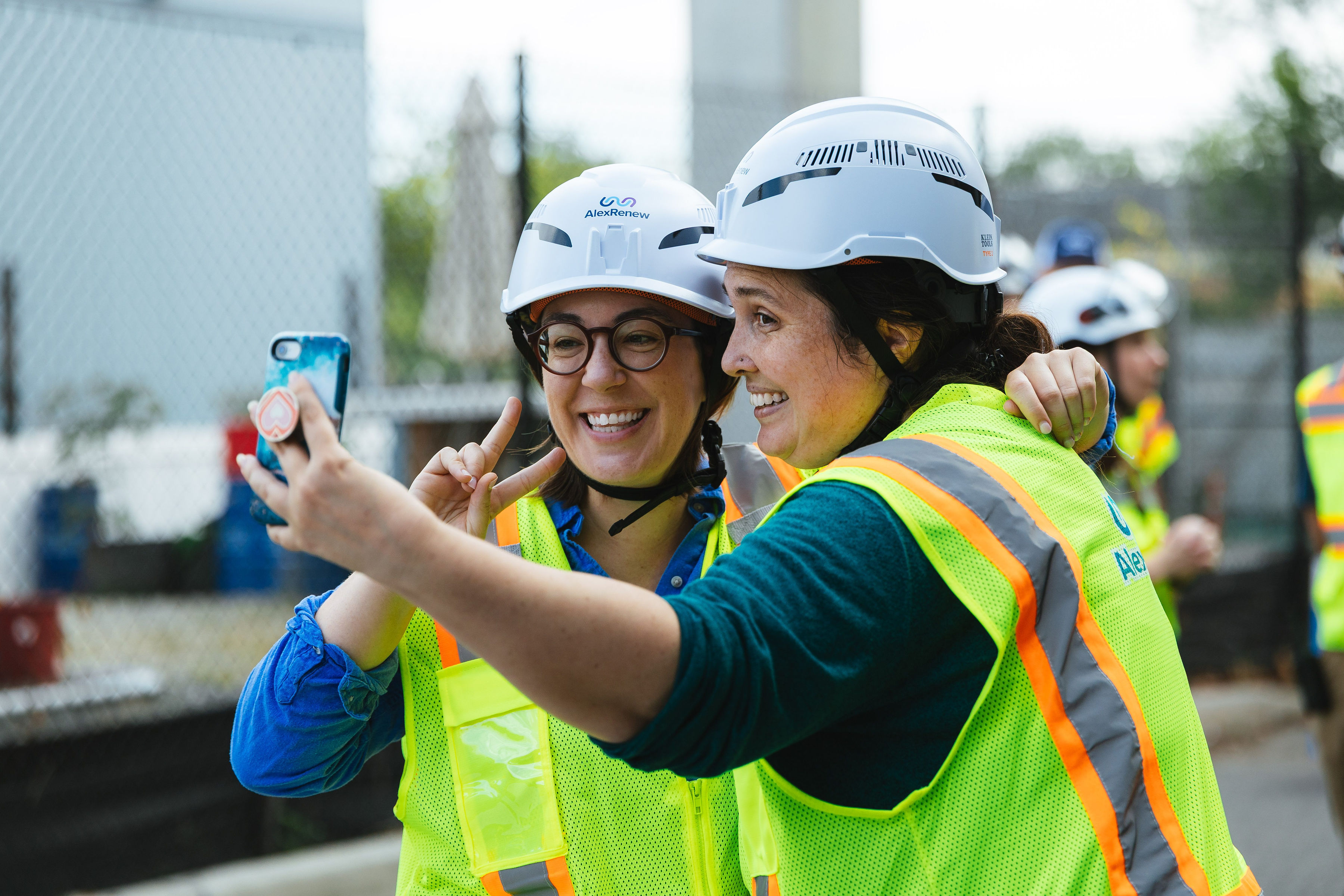 Two women in hard hats and safety vests smile at a cell phone camera.