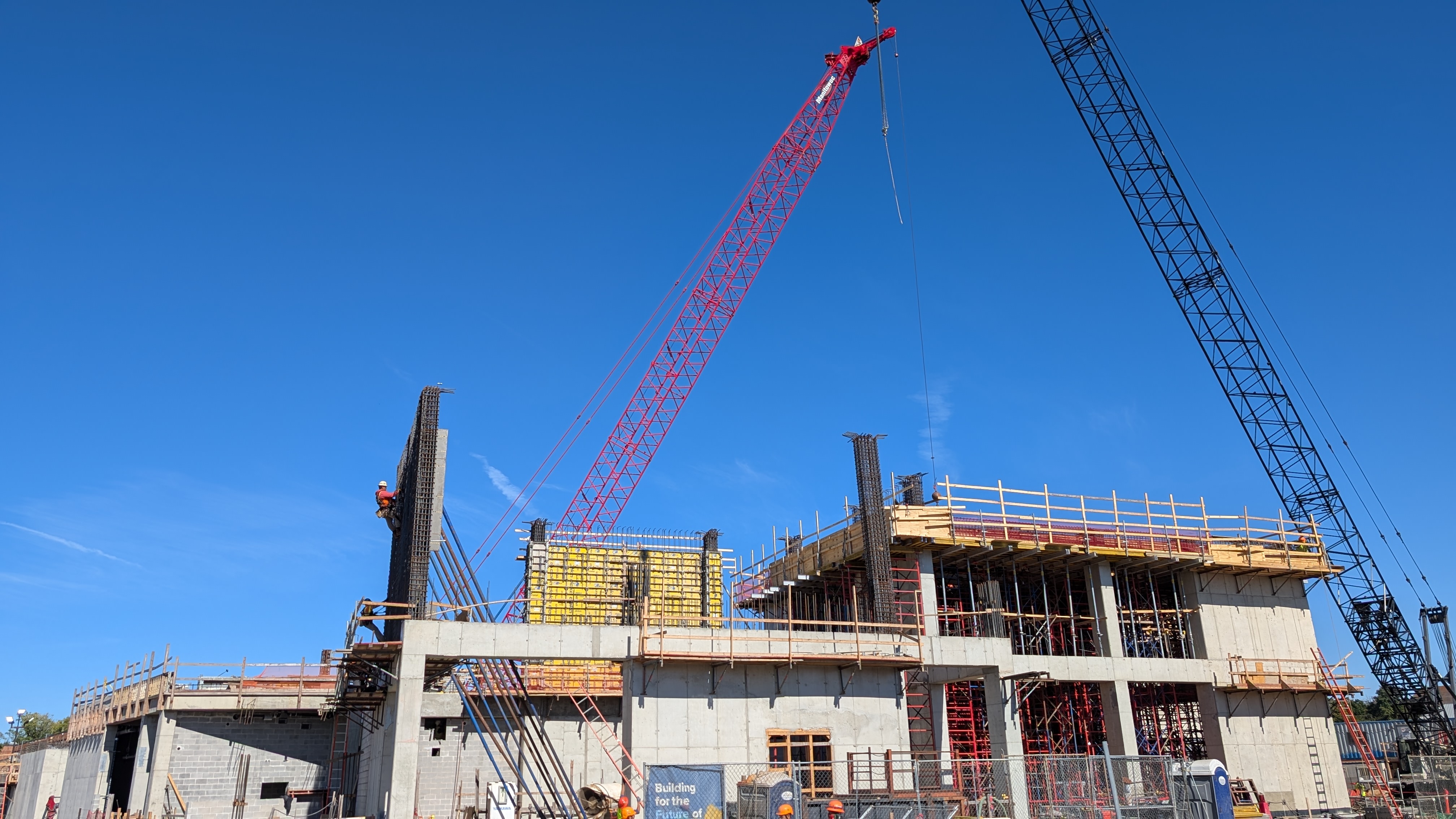 A half constructed building with two cranes towering above it. A blue sky is in the background.