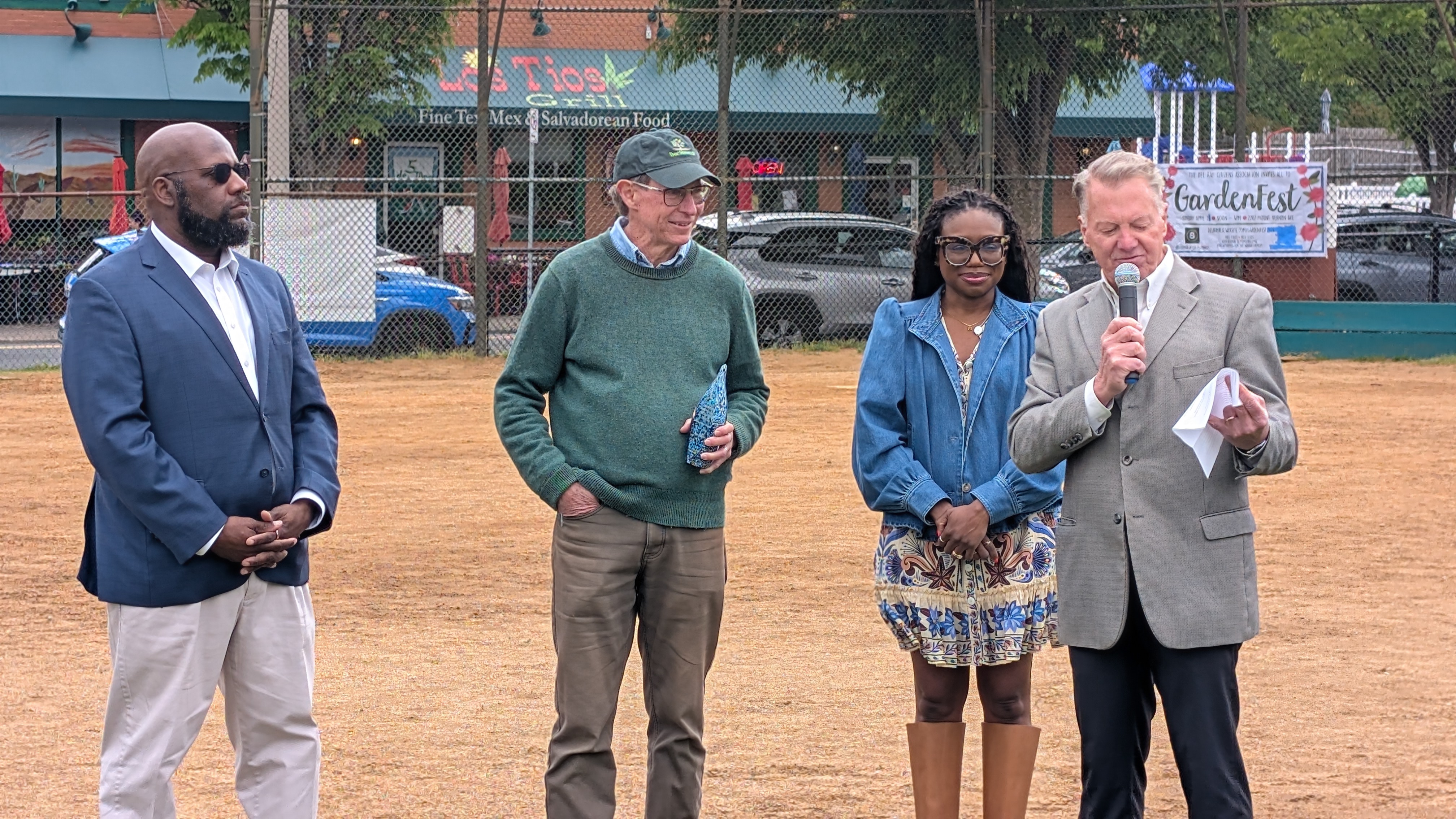 A man in a grey suit speaks into a microphone while three people look on.