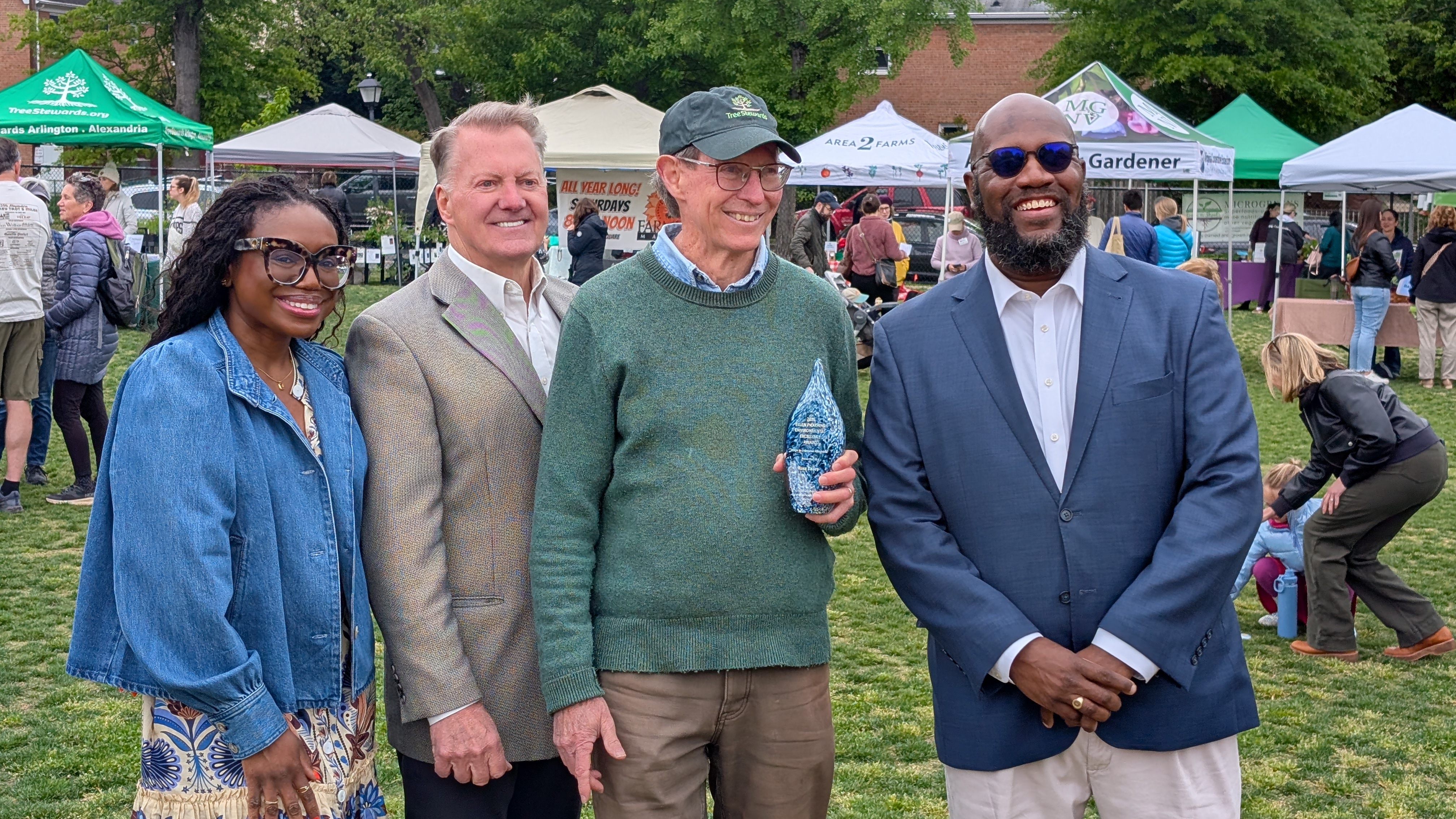Three people pose around a man wearing a green sweater and baseball cap who is holding an award.