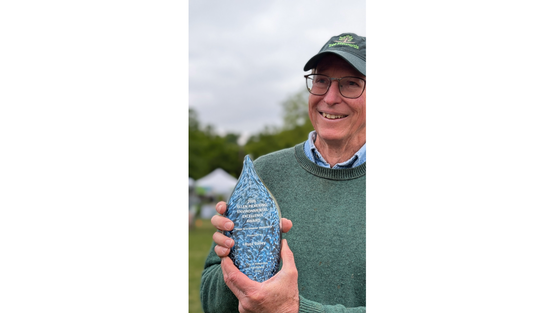 A man wearing a green sweater and baseball cap holds a blue award.