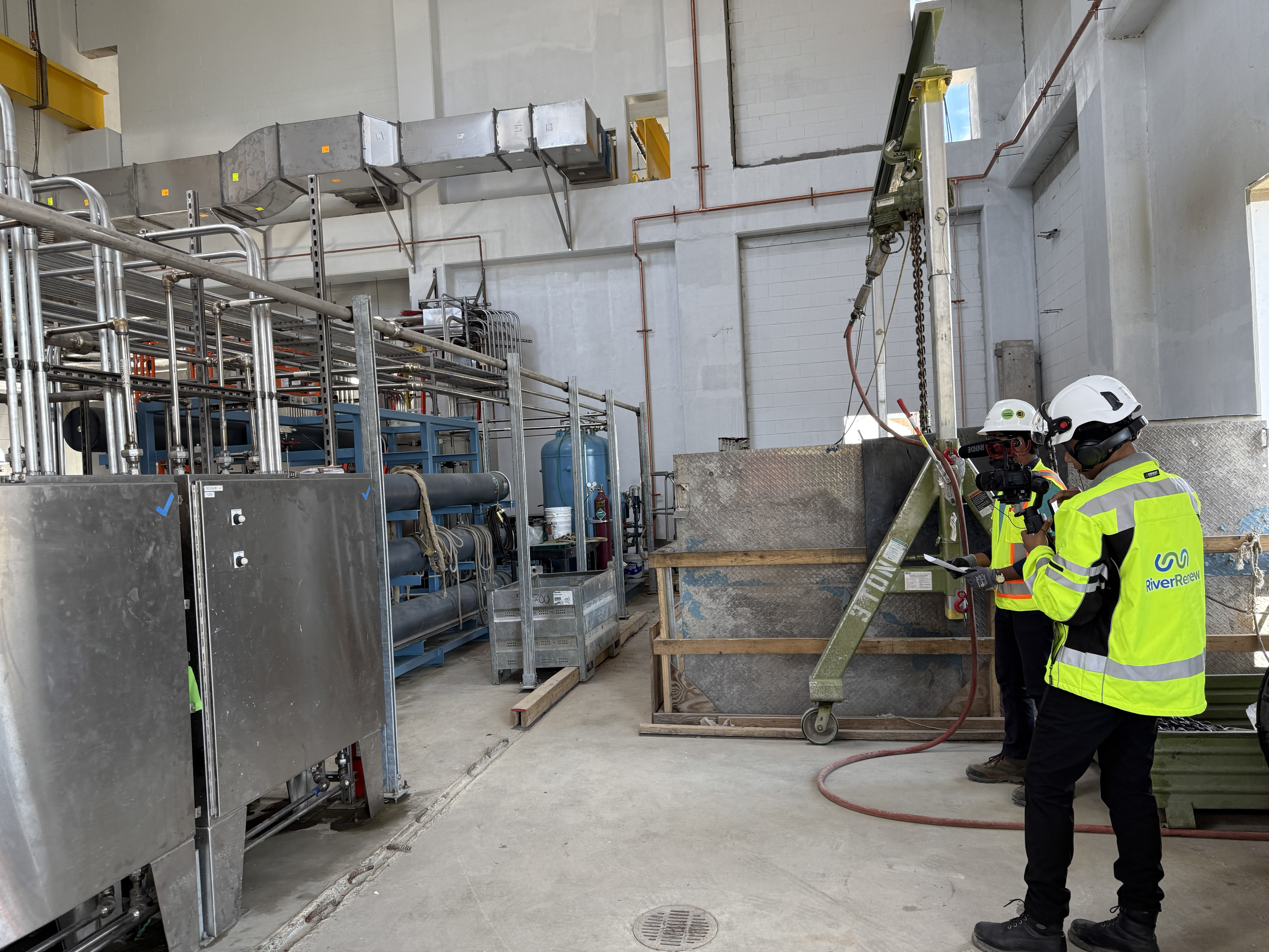 Two people in high vis safety gear examine a series of silver metal boxes and pipes inside a white-walled room.