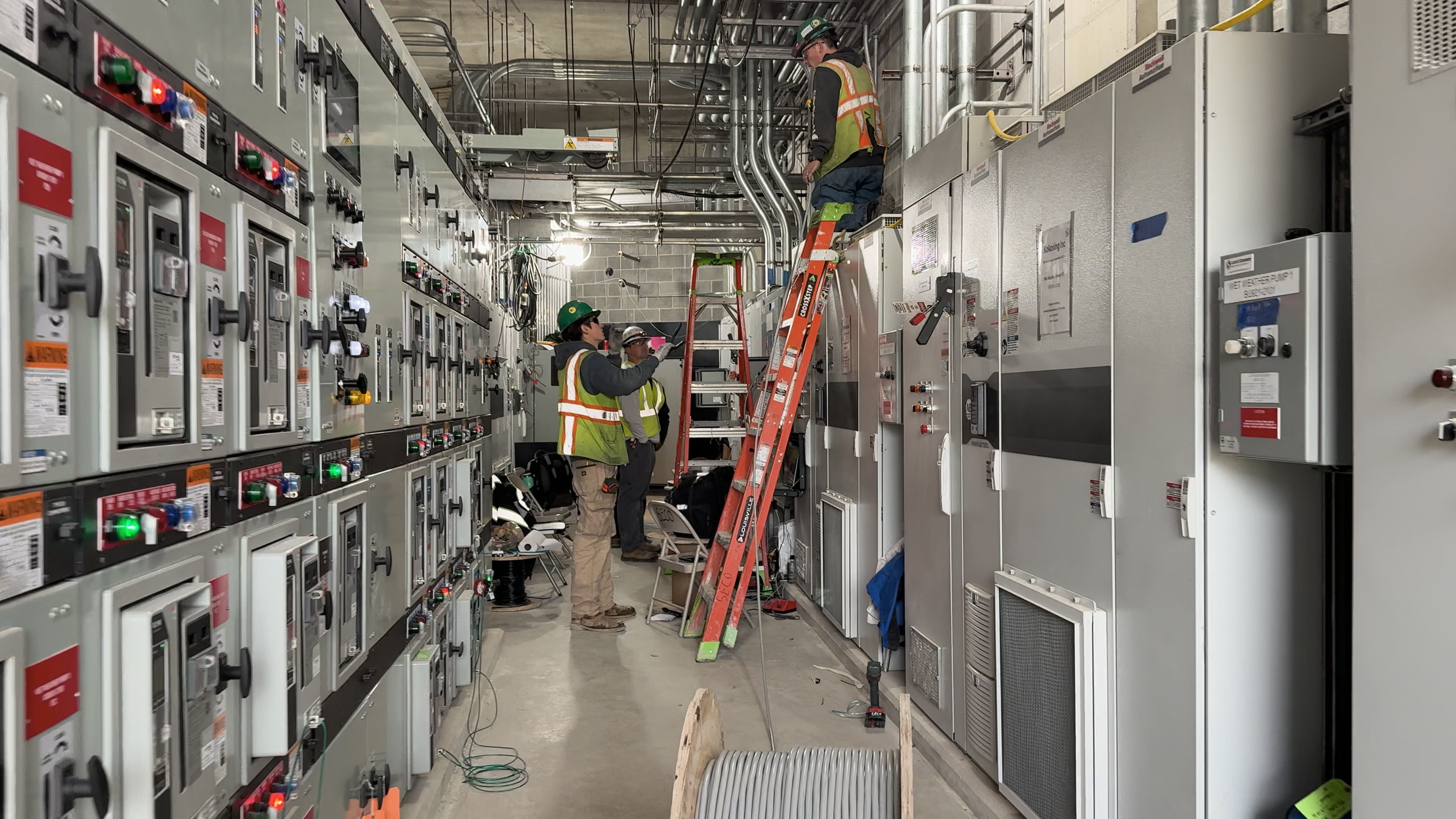 People in safety gear examine computers and electronics panels inside a hallway.