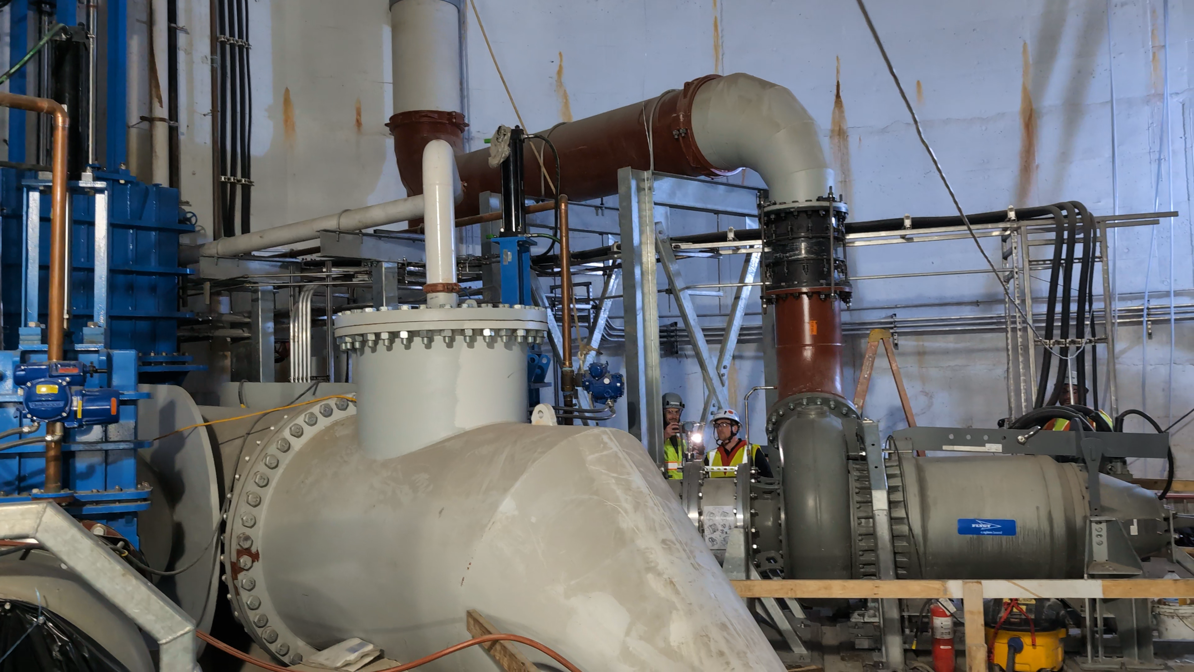 Large white pumps and red and blue pipes fill a high-ceilinged, underground room.