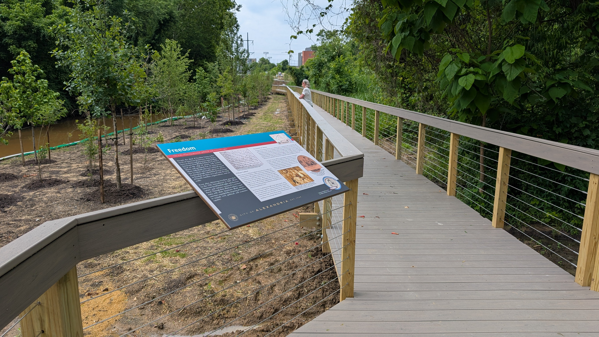 A new wayfinding sign installed on the railing of a boardwalk overlooks a newly restored riparian zone.