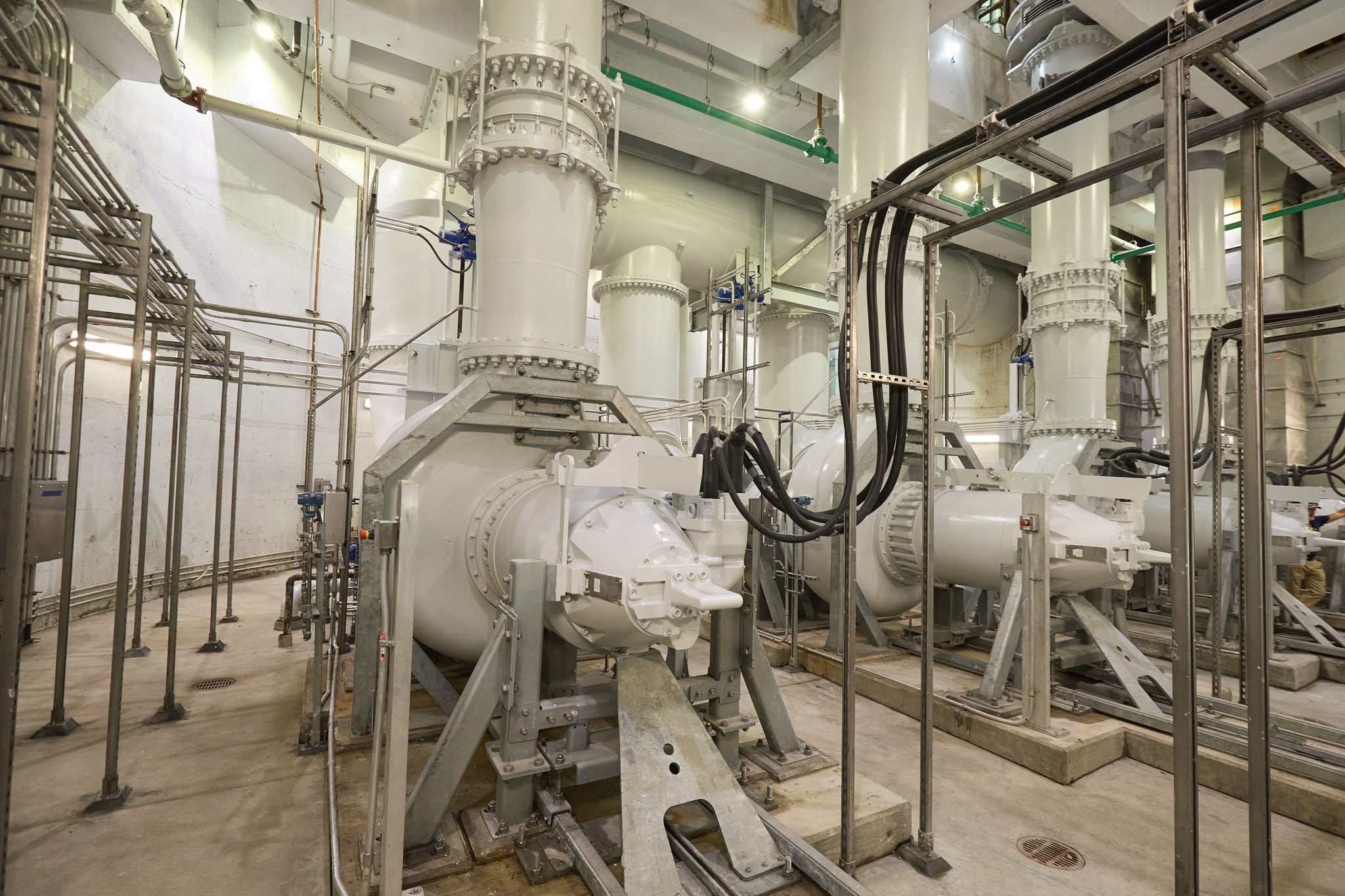 Large white pumps and pipes fill a high ceilinged room lit by florescent lights.
