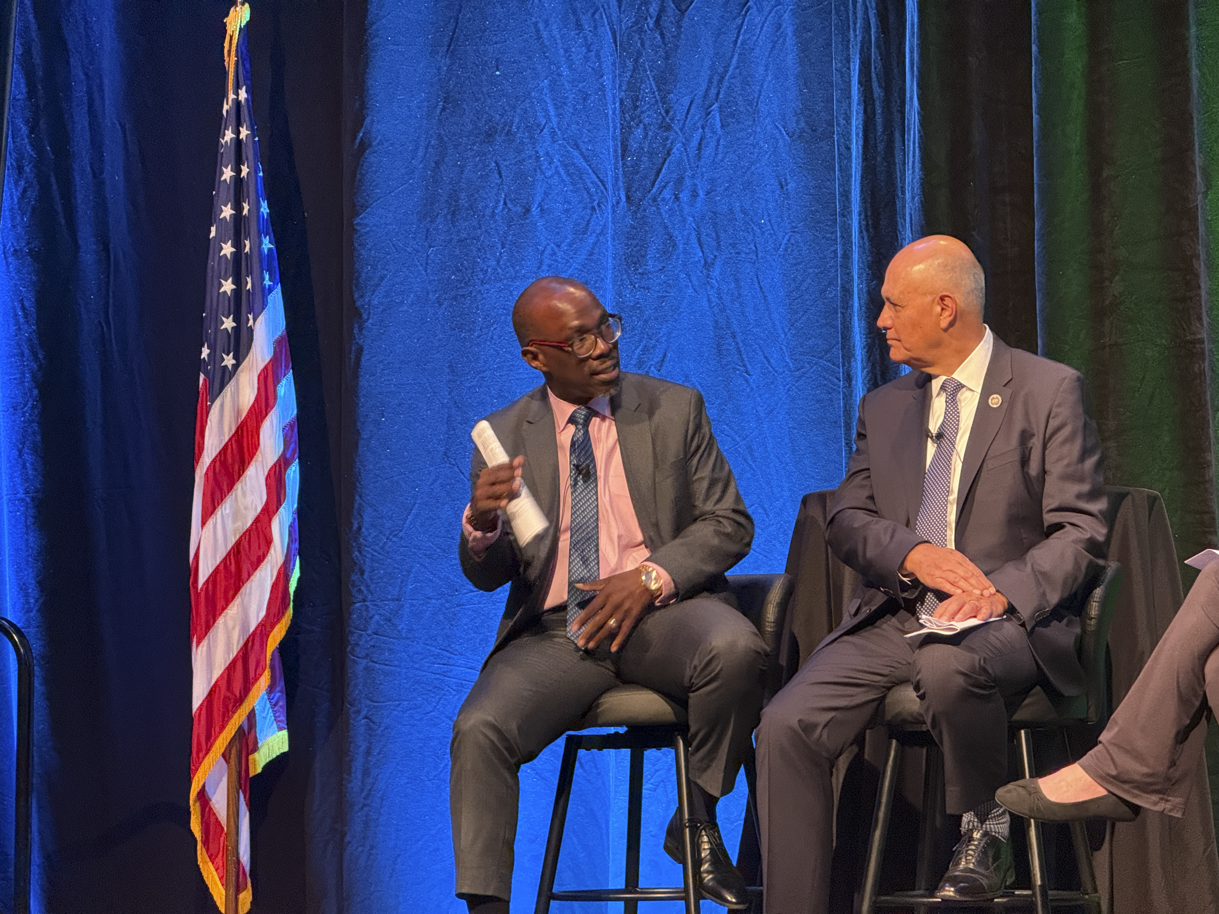 Two men on a conference panel speaking in front of an American flag.