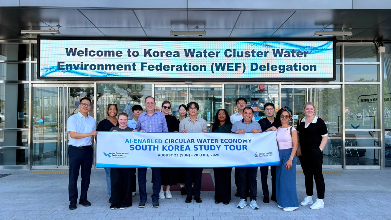 A group of people hold a banner beneath a sign that says "Welcome to Korea Water Cluster, Water Environment Federation (WEF)"