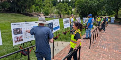 Staff in safety vests talk to visitors in front of a series of signs explaining the RiverRenew project. The signs are laid out in a line along a brick sidewalk.