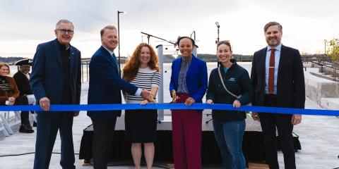 Six men and women in business attire stand behind a blue ribbon. The woman in the center, the mayor, holds a pair of scissors ready to cut the ribbon.