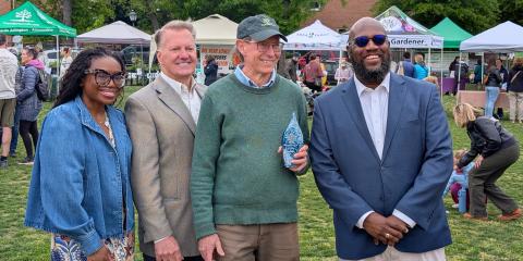 Three people pose around a man wearing a green sweater and baseball cap who is holding an award.