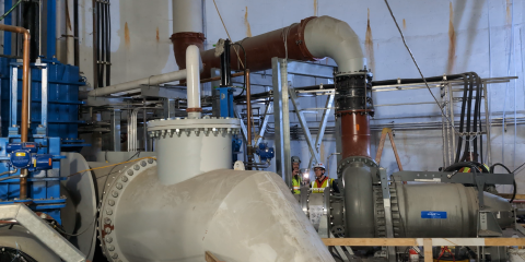 Large white pumps and red and blue pipes fill a high-ceilinged, underground room.