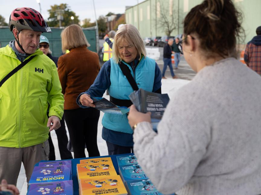 An elderly couple examine pamphlets at an AlexRenew informational table. An AlexRenew employee on the right side of the frame offers assistance.