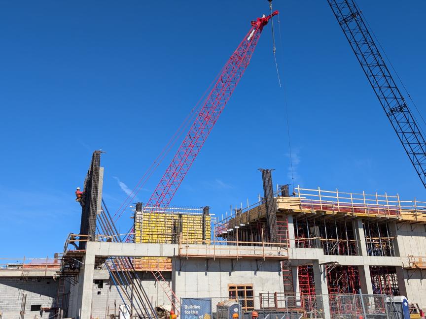 A half constructed building with two cranes towering above it. A blue sky is in the background.